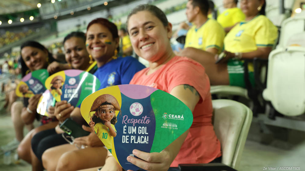 Sentadas lado a lado em cadeiras de uma arquibancada lotada de estádio, quatro mulheres sorridentes exibem, para a foto, leques da campanha.