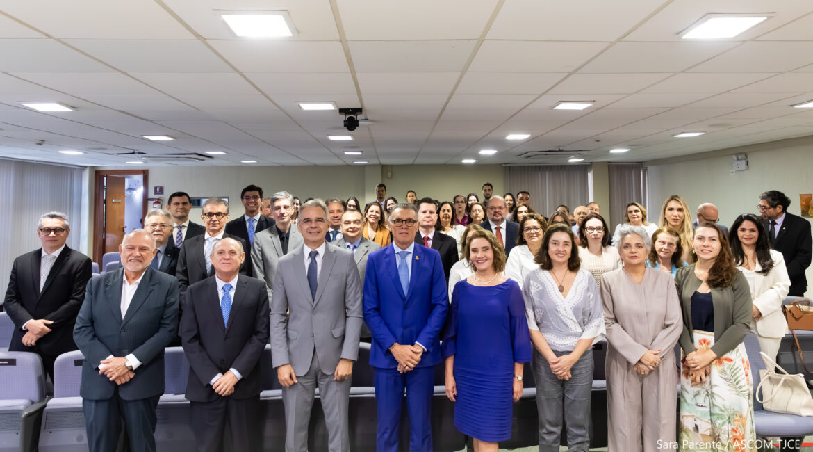 Imagem de várias pessoas posando para foto, sorrindo, em pé, em um grande auditório