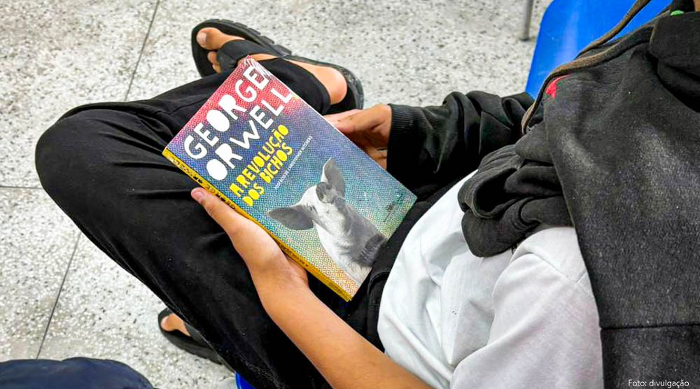Fotografia com a vista de cima de um estudante sentado com um livro nas mãos, apoiado na perna esquerda, que veste calça preta e sandália de dedo e está cruzada sobre a outra.