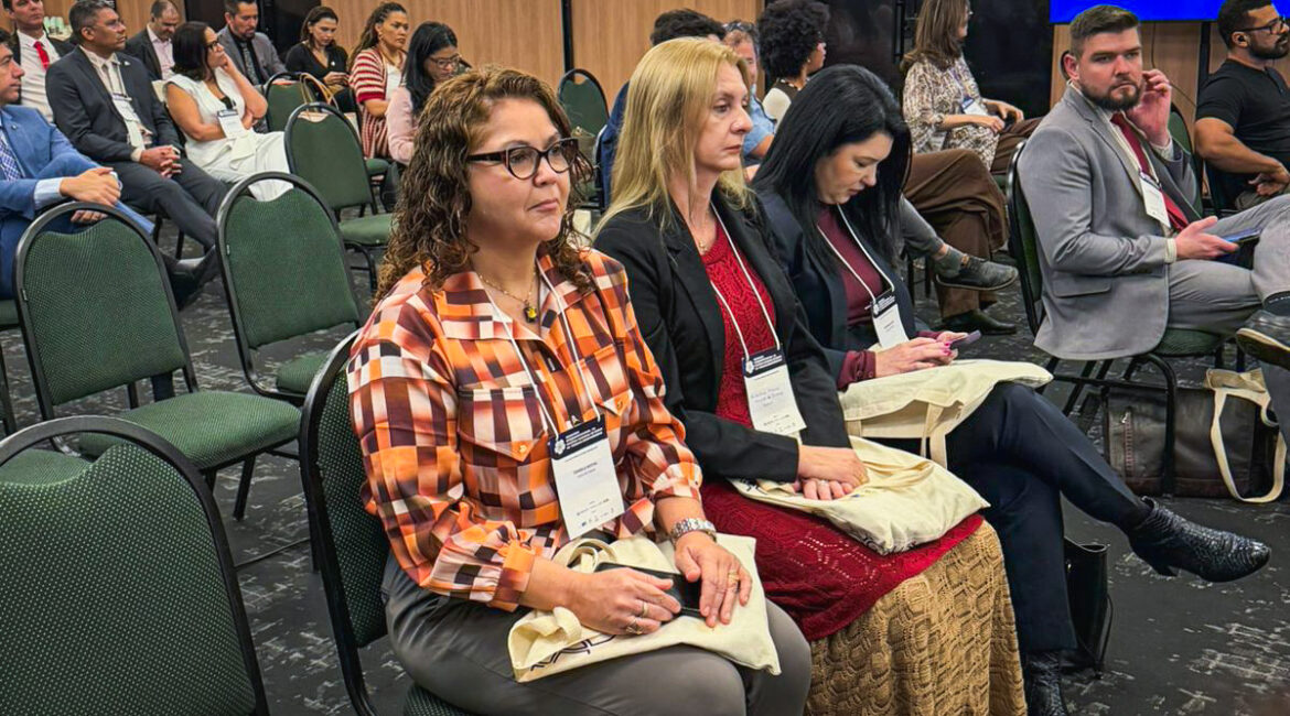 Foto de um auditório, com várias pessoas sentadas, com destaque para uma fila que tem três mulheres Foto de um auditório, com várias pessoas sentadas, com destaque para uma fila que tem três mulheres