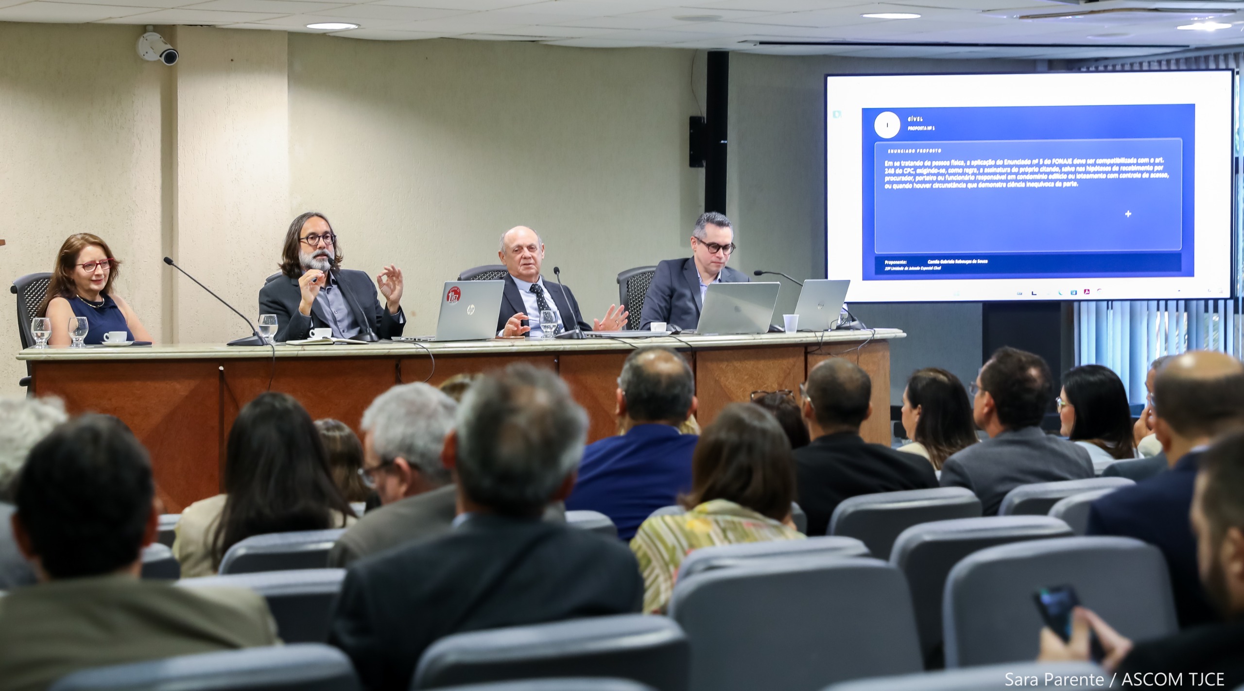 Imagem de uma mesa no palco na qual estão sentadas quatro pessoas. À frente delas, a plateia sentada em um grande auditório