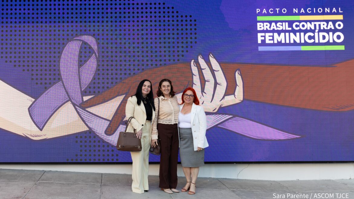 Foto de três mulheres posando para foto, sorrindo, em pé, em um amplo espaço cuja parede tem um simbolo que representa a luta contra o feminicídio no Brasil