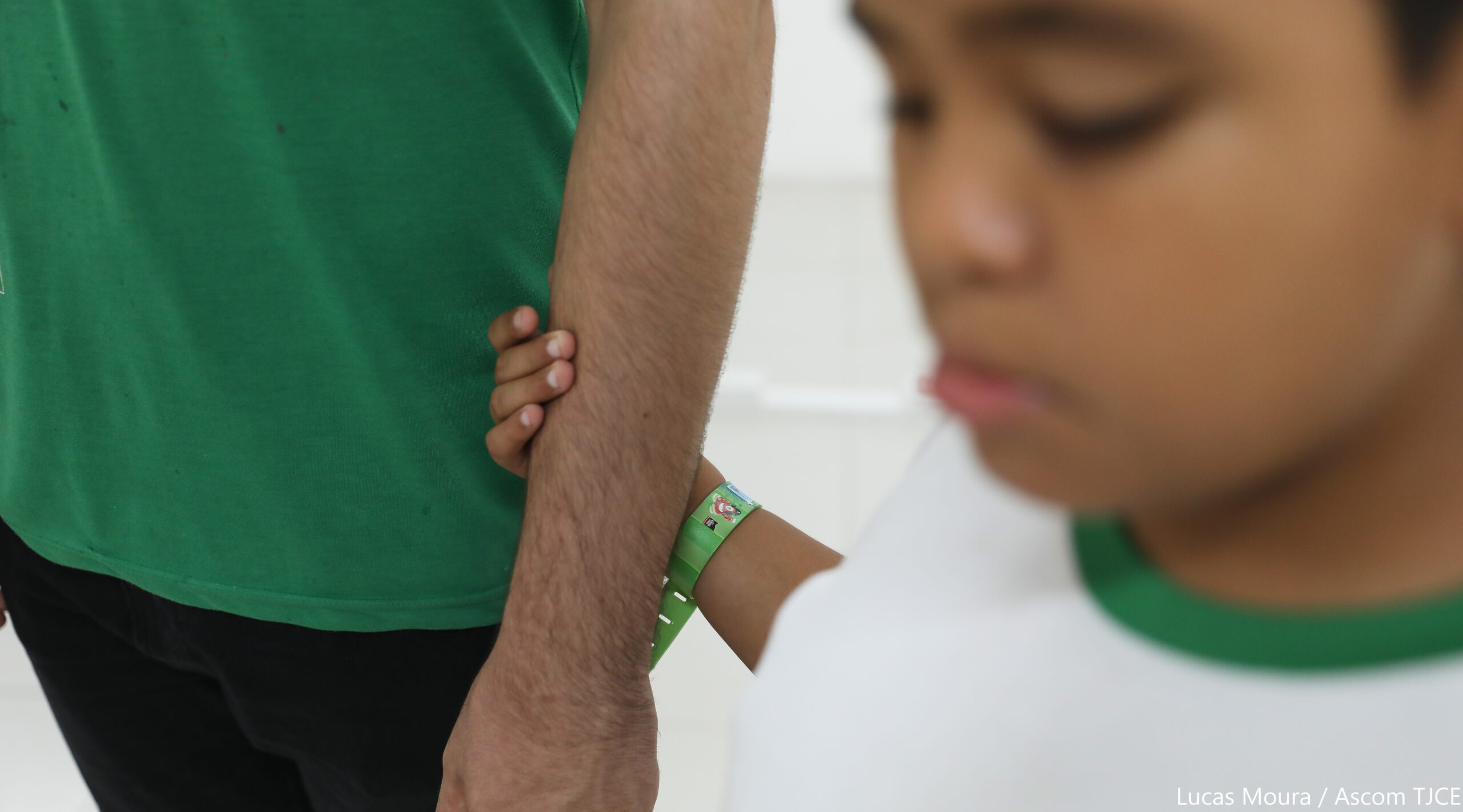 Imagem do rosto de um menino de 11 anos e da mão dele, segurando no braço de um adulto