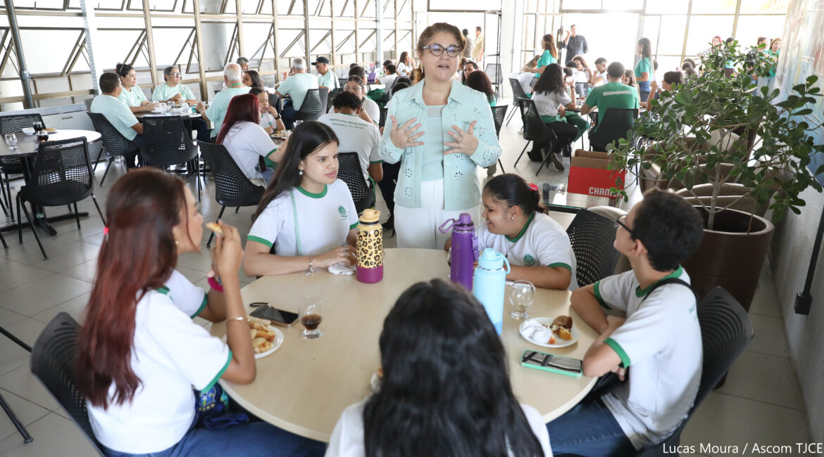 Foto de uma mulher de óculos, cabelos presos, em pé, falando próxima a cinco estudantes sentadas que estão lanchando