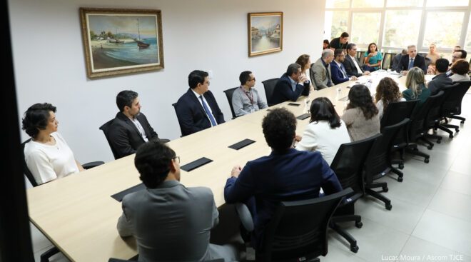 A imagem mostra muitas pessoas sentadas ao redor de uma grande mesa de reuniões