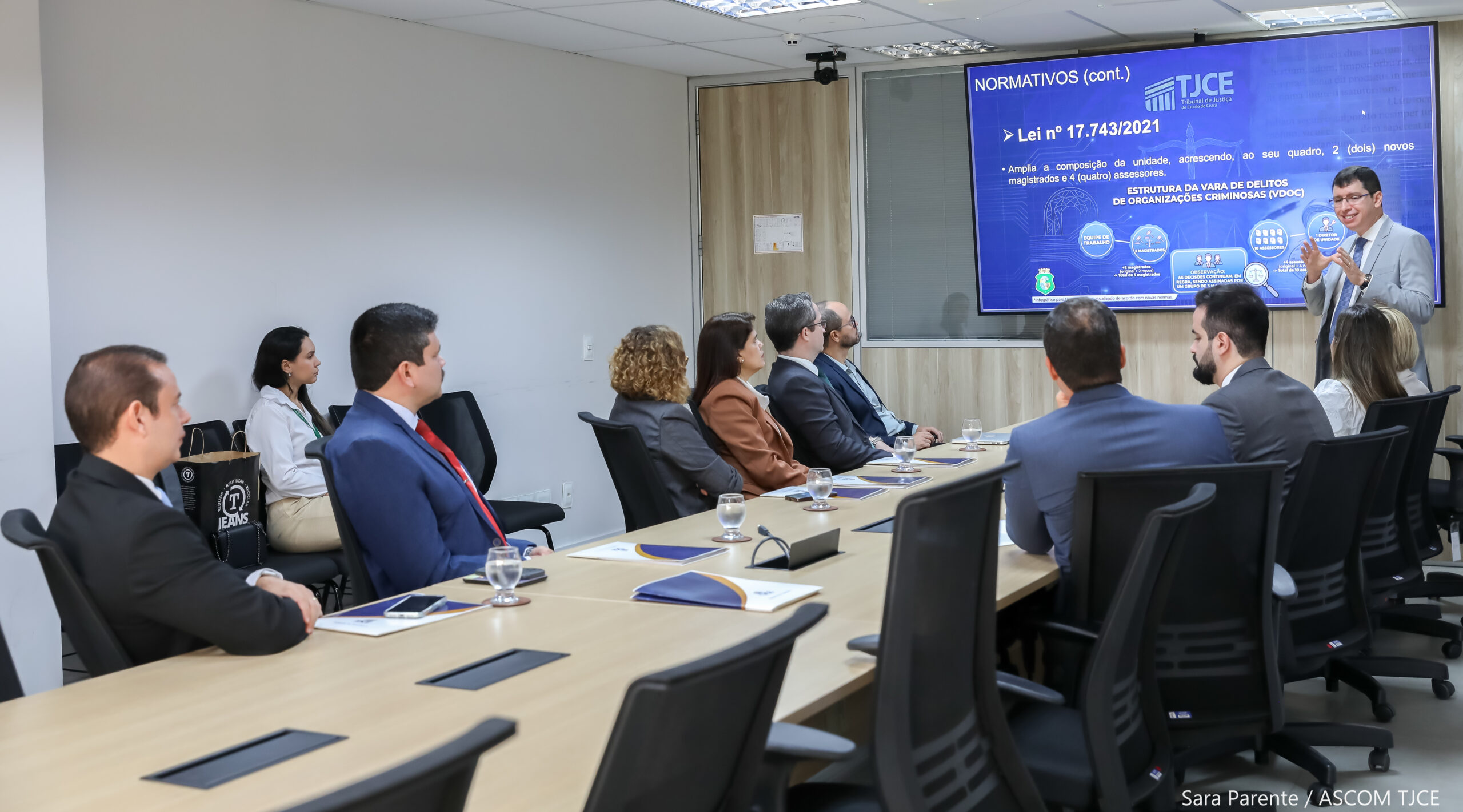 Imagem de várias pessoas sentadas em uma mesa de trabalho, atentas às explicações de um homem à frente deles, falando das informações que estão projetadas em grande tela na prarede