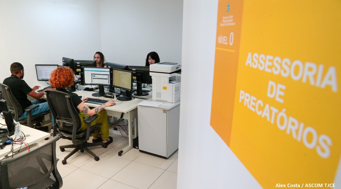 Foto de quatro pessoas sentadas numa bancada de trabalho e, à frente delas, na parede, um banner na parede escrito Assessoria de Precatórios