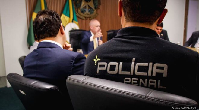A imagem mostra as costas de um homem cuja camisa identifica Polícia Penal. Ao redor dele, várias pessoas sentadas em reunião