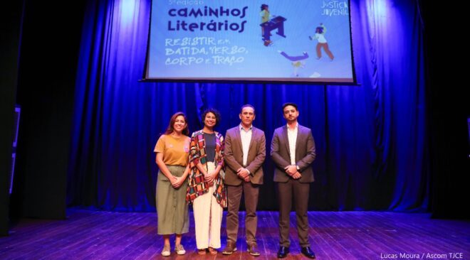 Duas mulheres e dois homens de frente em um palco com um grande quadro azul ao fundo escrito "Caminhos Literários"