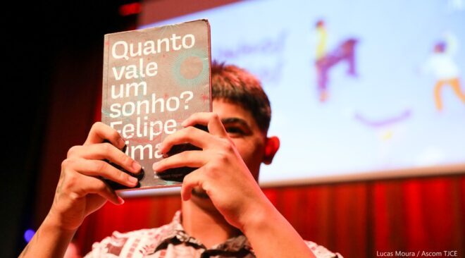 Homem jovem seguro um livro com o título "Quato vale um sonho" em frente ao rosto. Ao fundo tem um grande quadro azulado com imagens desfocadas