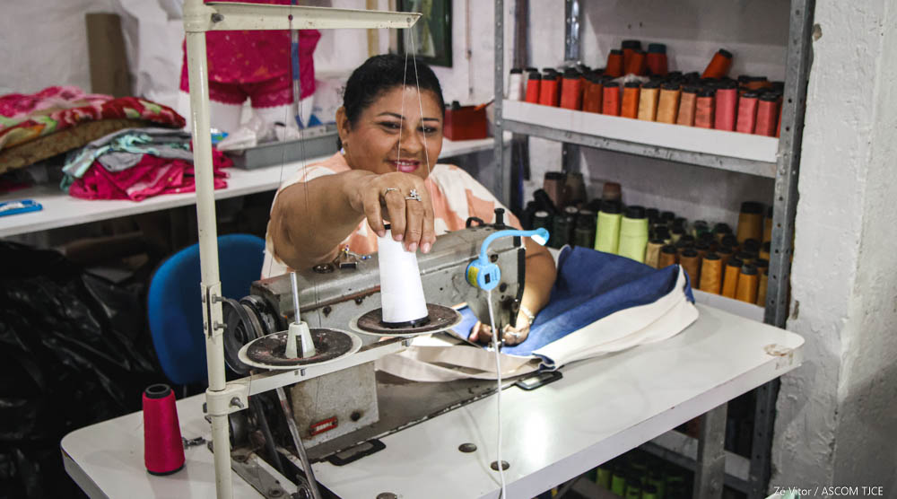 Mulher sentada numa mesa de costura, que uma máquina. E ela está segurando um tubo de linha, sorrindo para foto.