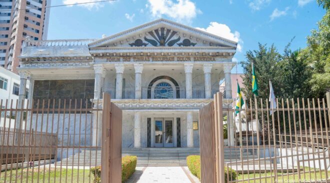 Prédio com térreo e andar superior em arquitetura clássica. Ao lado do edifício as bandeiras do Estado, Brasil e Poder Judiciário