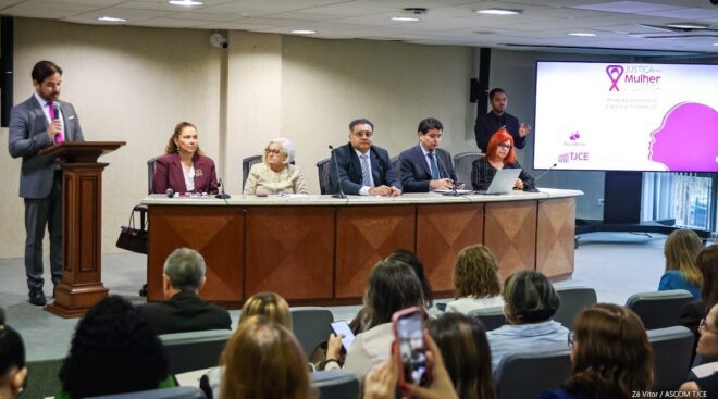 Cinco pessoas vestidas formalmente prestam atenção a um homem que está em pé lendo, Ao fundo, outro homem interpreta em libras o que está sendo dito