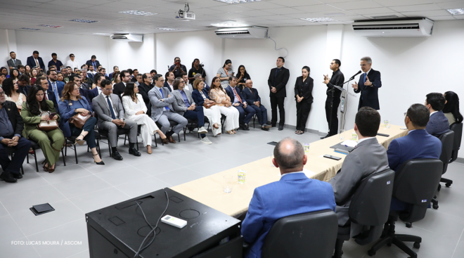 Homem com microfone discursa para muitas pessoas sentadas em uma sala ampla