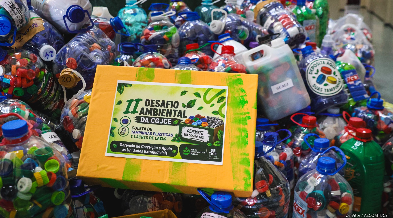 Mês da Sustentabilidade 1-1783363032 Caixa amarela com os dizeres Segundo Desafio Ambiental. Ao redor da caixa diversas garrafas plásticas cheias de tampinhas coloridas.