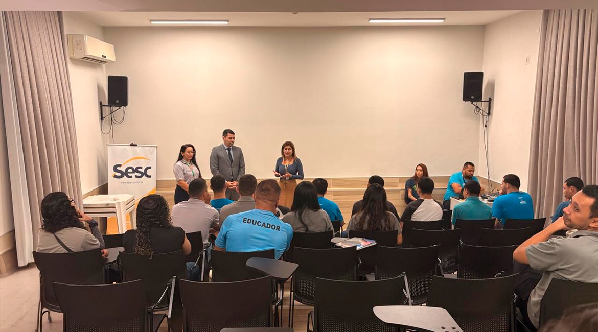 Imagem de um auditório em que estão pessoas em pé, na frente, duas mulheres e um homem, falando para um grupo de jovens que estão sentados