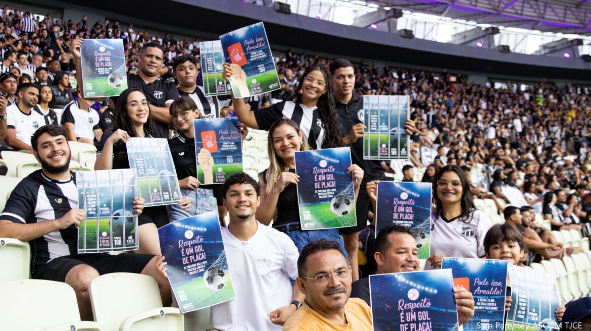 Várias pessoas sentadas na arquibancada do estádio de futebol mostrando cartaz, na cor azul e verde, onde se lê: Respeito é um gol de placa