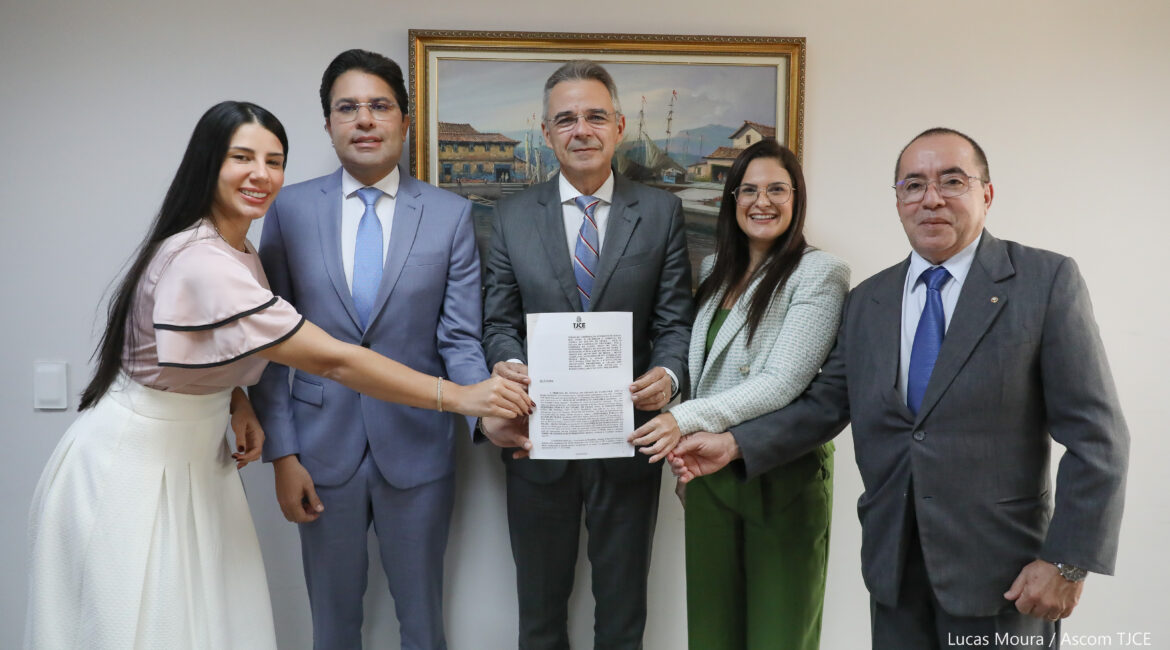 Imagem de três homens e duas mulheres posando para foto, sorrindo. Todos seguram uma cópia do termo de adesão