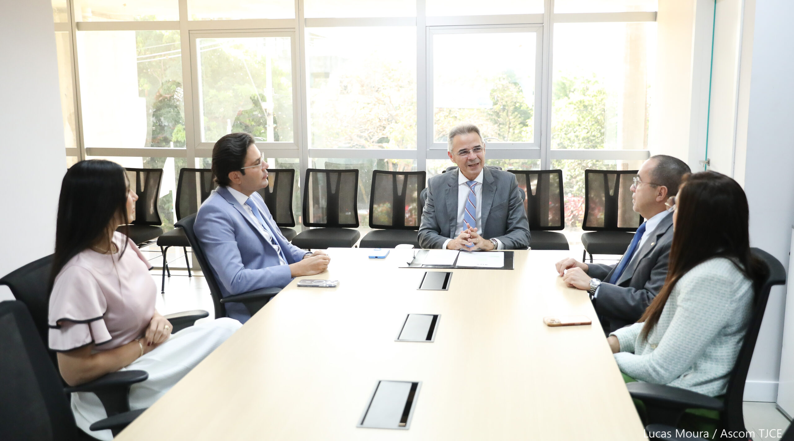 Imagem de cinco pessoas sentadas em uma mesa de trabalho, dialogando