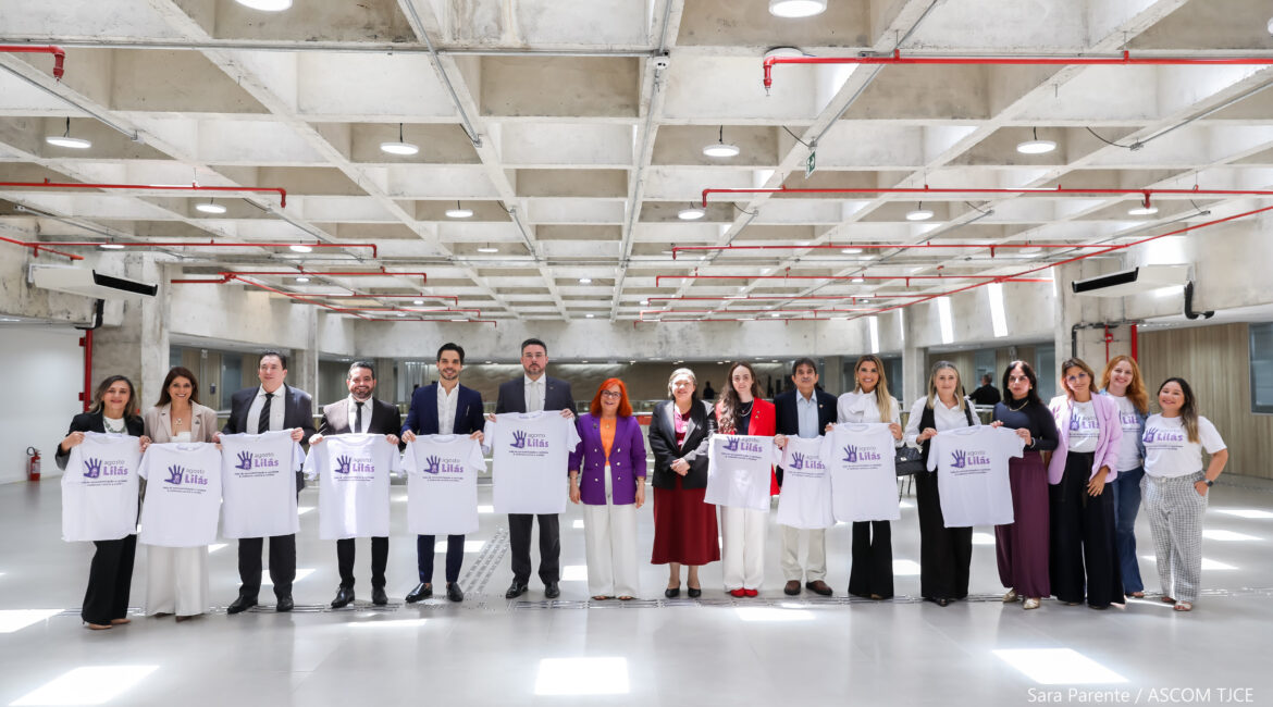 Várias mulheres e quatro homens posando para foto, segurando a blusa da campanha Agosto Lilás