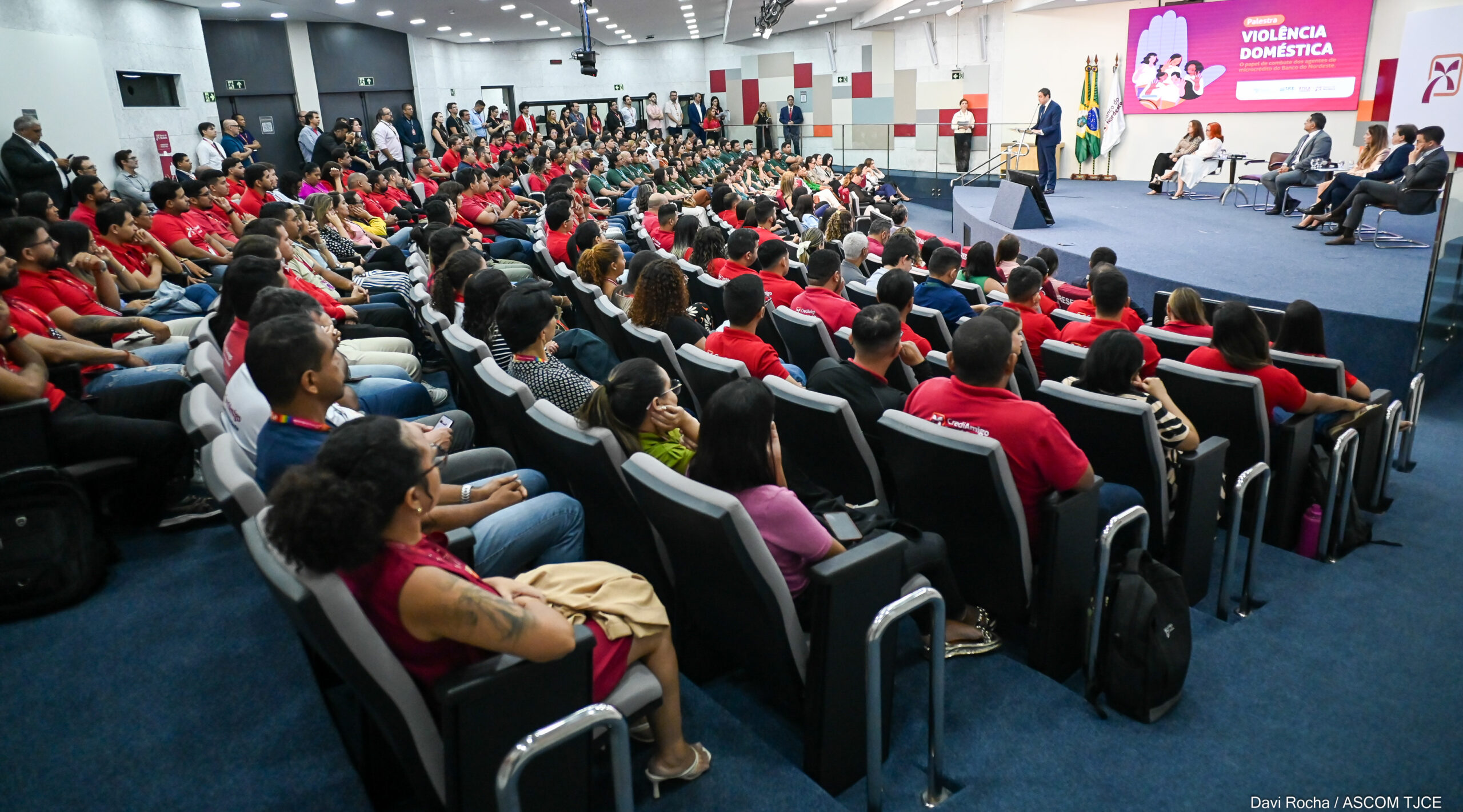 Foto panorâmica do evento, com várias pessoas sentadas na plateia e, à frente delas, pessoas sentadas no palco e um pessoa falando ao microfone no púlpito