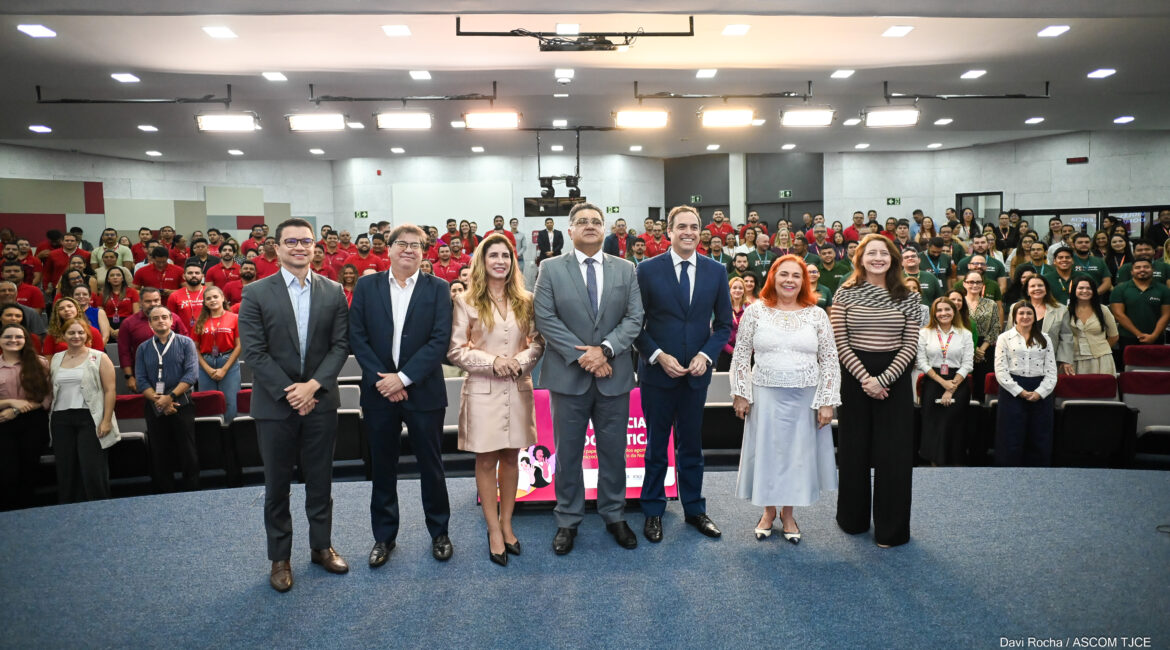 Foto de uma filha de homens e mulheres em destaque, posando para foto no palco em pé, e atrés deles, um grande número de pessoas no auditório também em pé