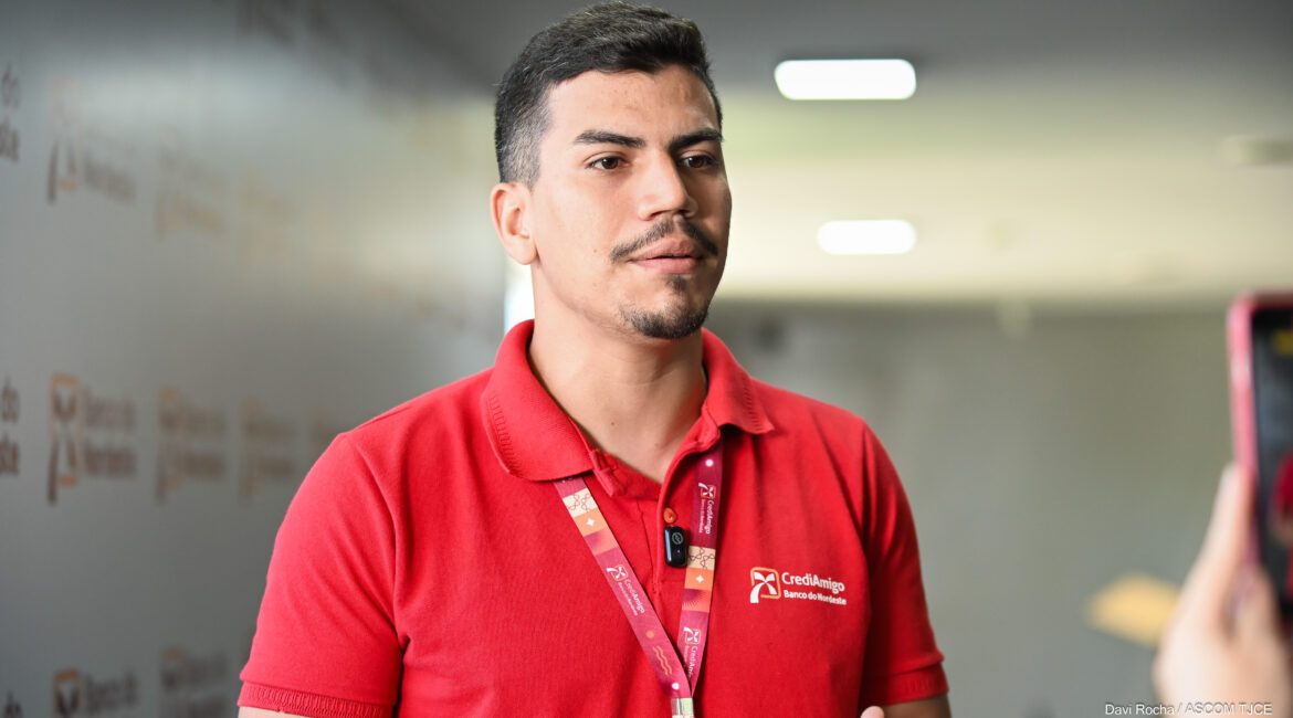 Foto de um rapaz em pé, vestido de blusa na cor vermelha, dando entrevista