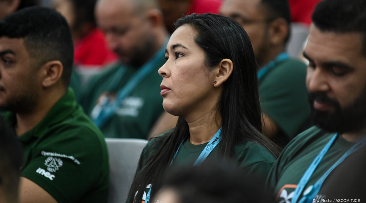 Foto de uma mulher em destaque, sentada, ladeada por dois homens também sentados. Todos estão vestidos de com blusas verdes