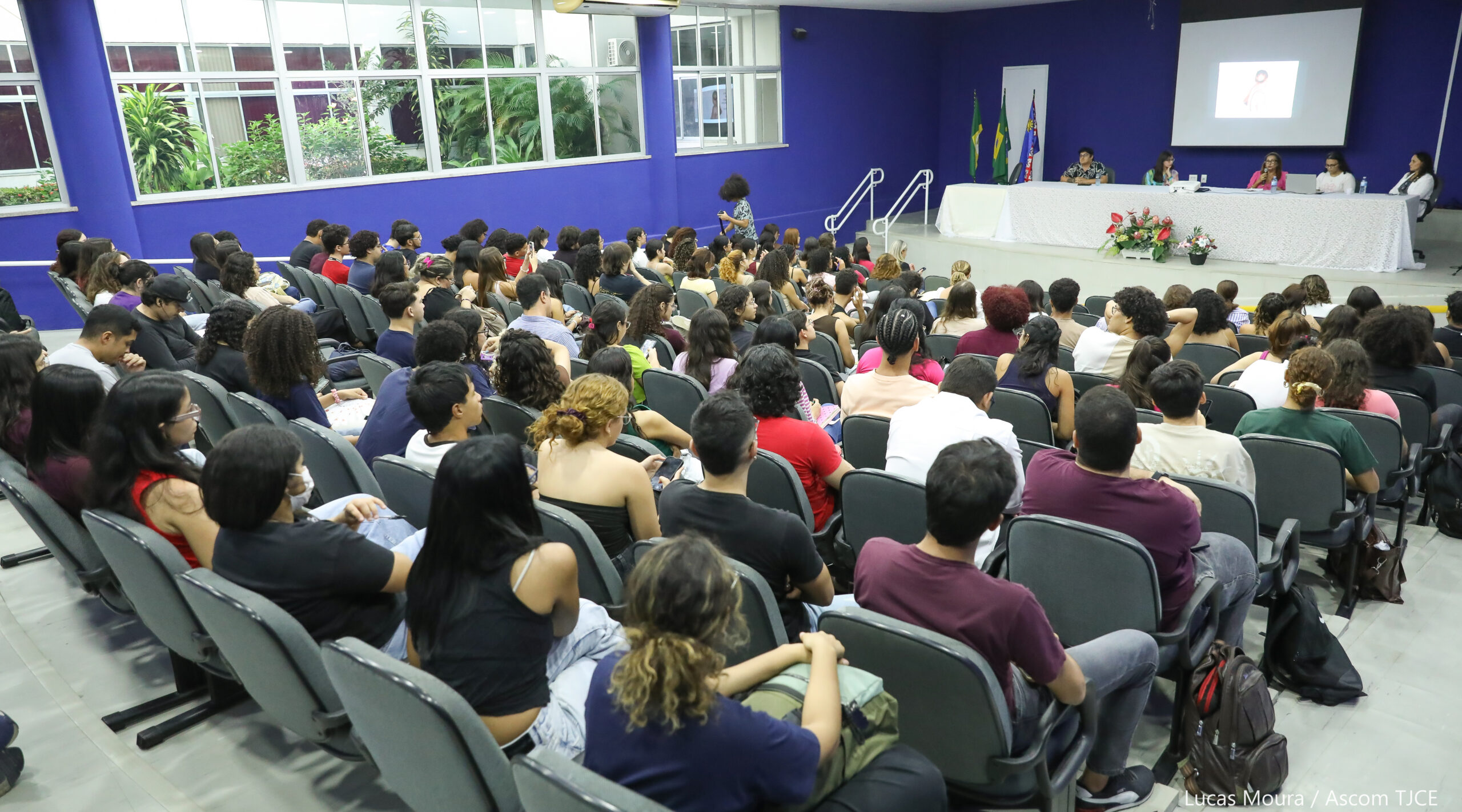Foto panorâmica do auditório com várias pessoas sentadas na plateia, e uma mesa no palco com mulheres e homens palestrando
