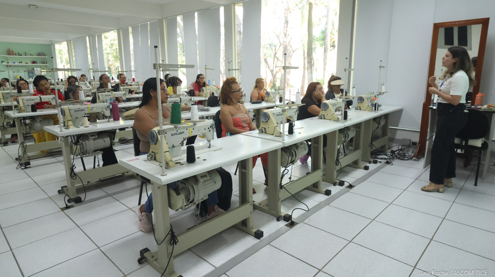 Imagem de uma sala mostrando várias mulheres sentadas, aprendendo a costurar. À frente delas, uma instrutora dando as explicações.
