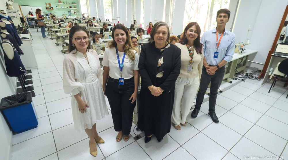 Imagem de quatro mulheres sorrindo para foto, em pé, em uma sala, além de um homem. Atrás delas, várias mulheres aprendendo a costura em máquinas de costurar