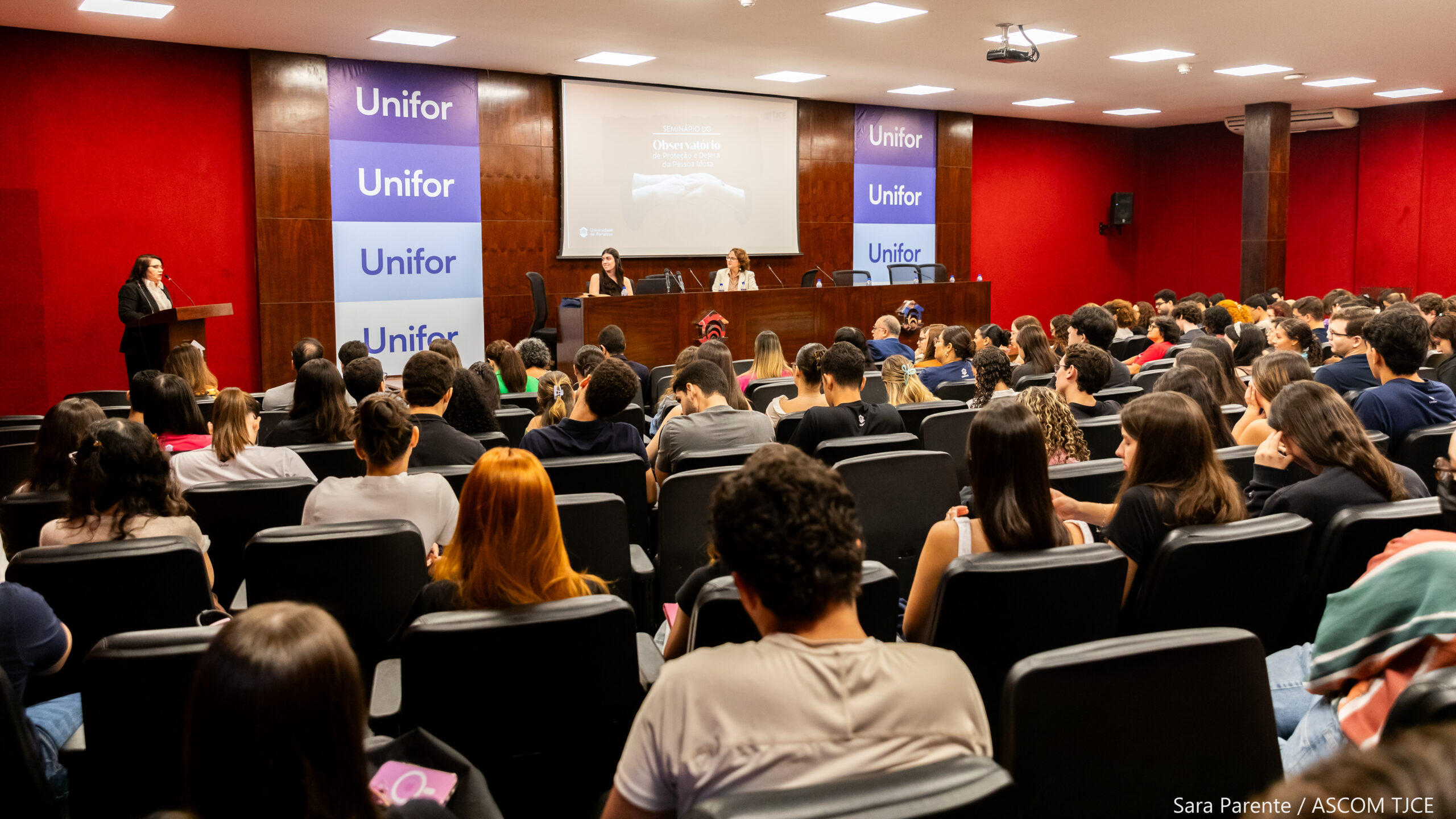 Imagem de um grande auditório lotado de pessoas sentadas. À esquerda, uma mulher no púlpito falando ao microfone.
