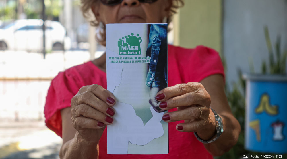 Imagem de uma mulher com destaque para o folder que ela segura nas mãos, onde se lê: Mães em luto!