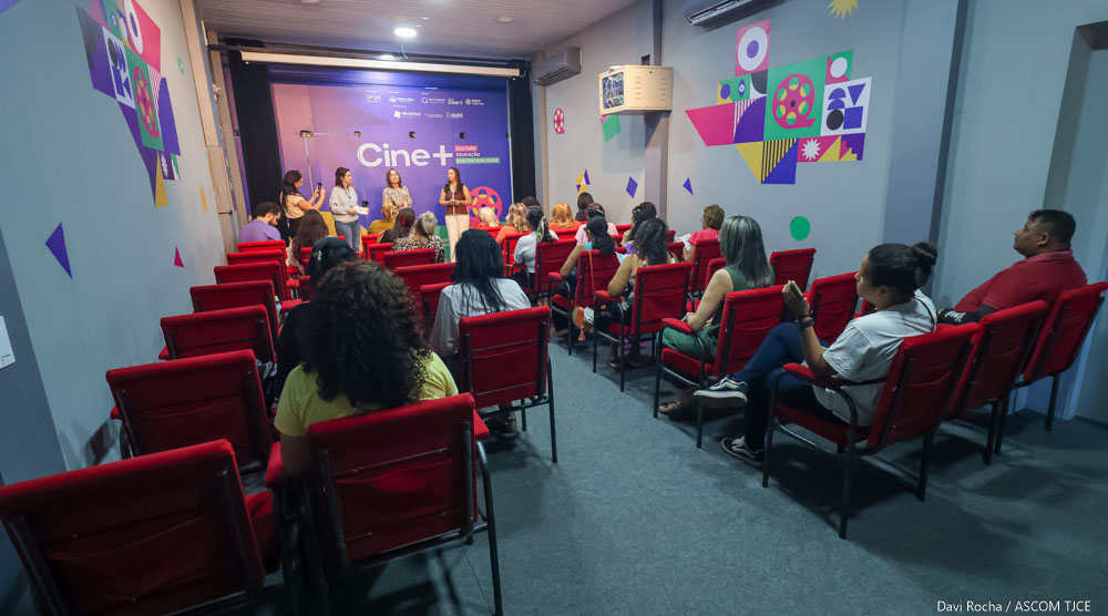 Imagem de várias pessoas sentadas em uma sala, ouvindo a mensagem de quatro mulheres que estão à frente delas