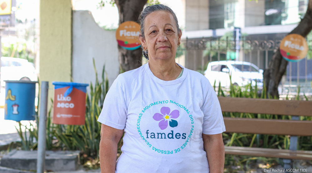 Imagem de uma mulher em pé, com uma blusa branca com o símbolo da entidade que ela fundou: Famdes