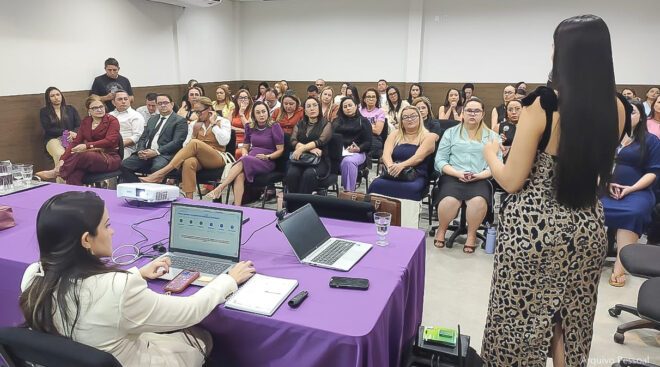 Mulher palsetrando para plateia em uma sala, ao lado de uma mesa com notebook.