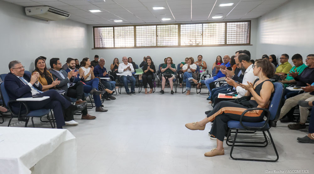 Imagem geral de uma sala ampla, com várias pessoas sentadas, batendo palmas