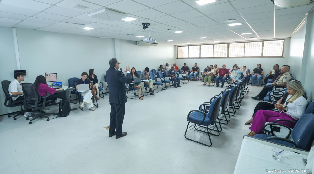 Foto de um homem em uma grande sala, falando em pé, ao microfone, e ao redor dele, várias pessoas sentadas, ouvindo-o