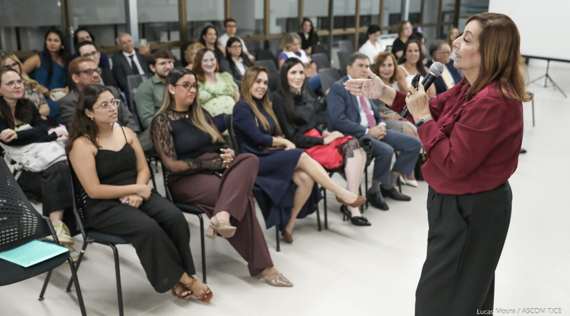 Mulher em pé, falando ao microfone e, à frente dela, várias pessoas sentadas na plateia ouvindo-a