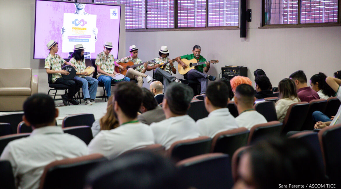 Seis homens no palco tocando instrumentos musicais, sendo cinco com chapus e um sem. À frente deles, a plateia sentada ouvindo