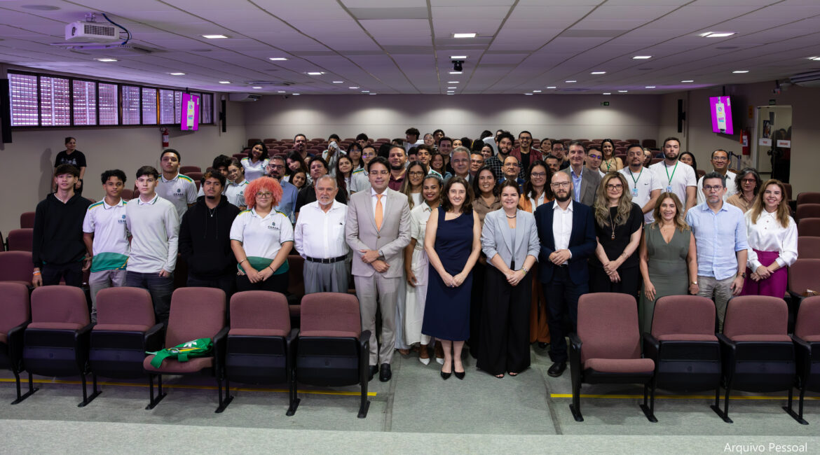 Imagem com várias pessoas posando para foto em pé, sorrindo, em um grande auditório