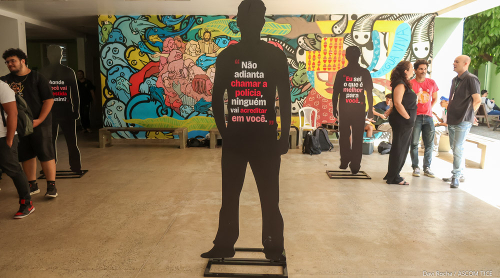 Fotografia de pessoas visitando a exposição destacando, ao centro, um dos totens de silhuetas masculinas em tamanho real, pretas e com frases no meio.