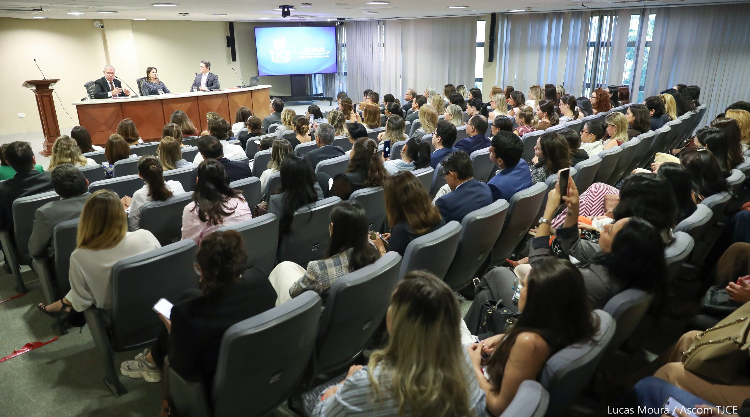 Foto geral de um auditório com várias pessoas sentadas e, à frente delas, dois homens e uma mulher sentados em uma mesa no palco, sendo que um dos homens fala ao microfone