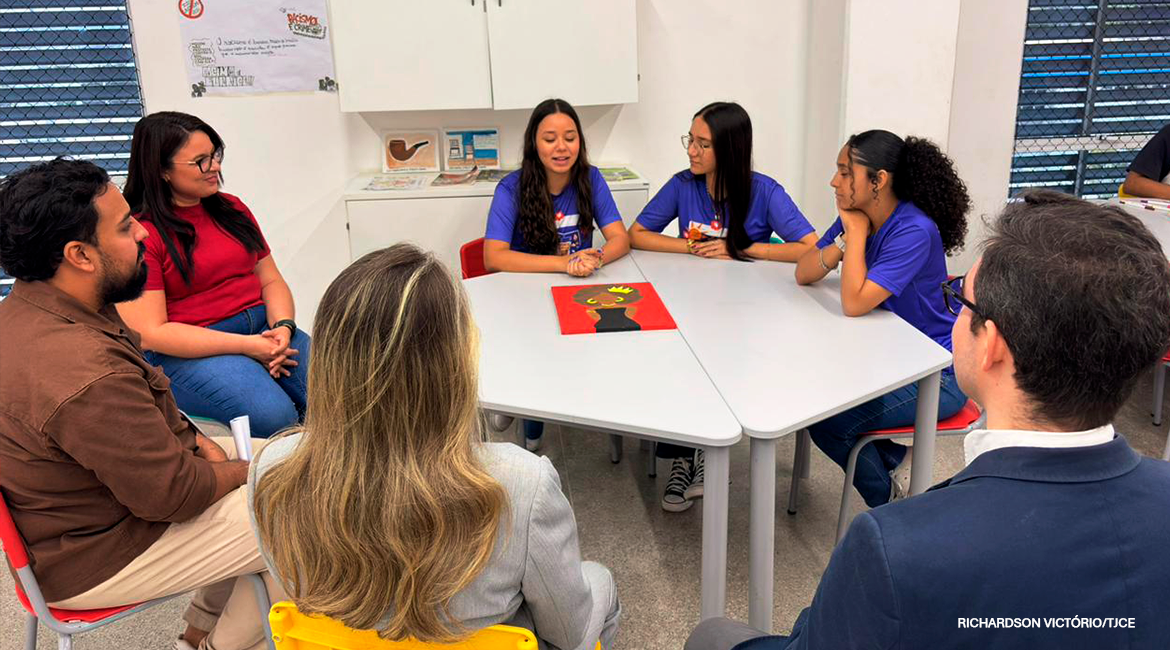 Foto de uma mesa em que se encontram três alunas, com professores do colégio e a juíza e o juiz do tribunal. Eles estão dialogando