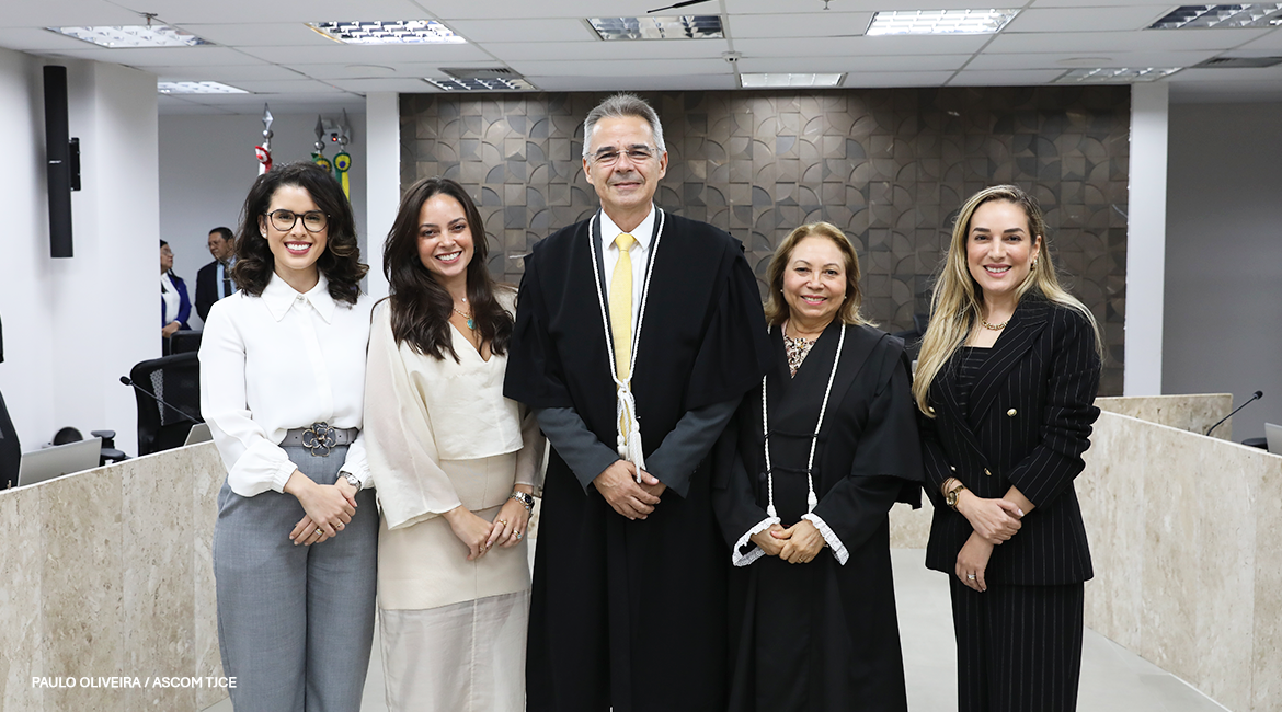 Foto de um homem e quatro mulheres, sorrindo para foto, em pé, em um salão
