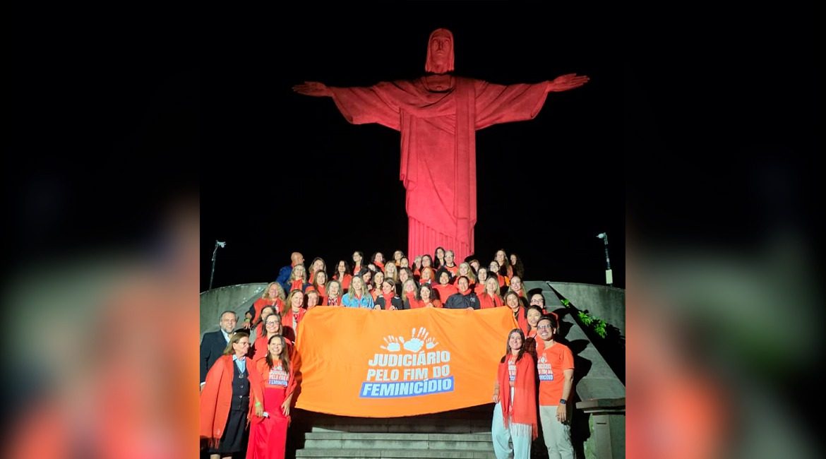 Imagem de várias pessoas à frente da fachada do Cristo Redentor no Rio de Janeiro, sorrindo para foto. Ao centro, todos segurando uma bandeira, nela inscrita: Pelo fim do feminicídio no Brasil