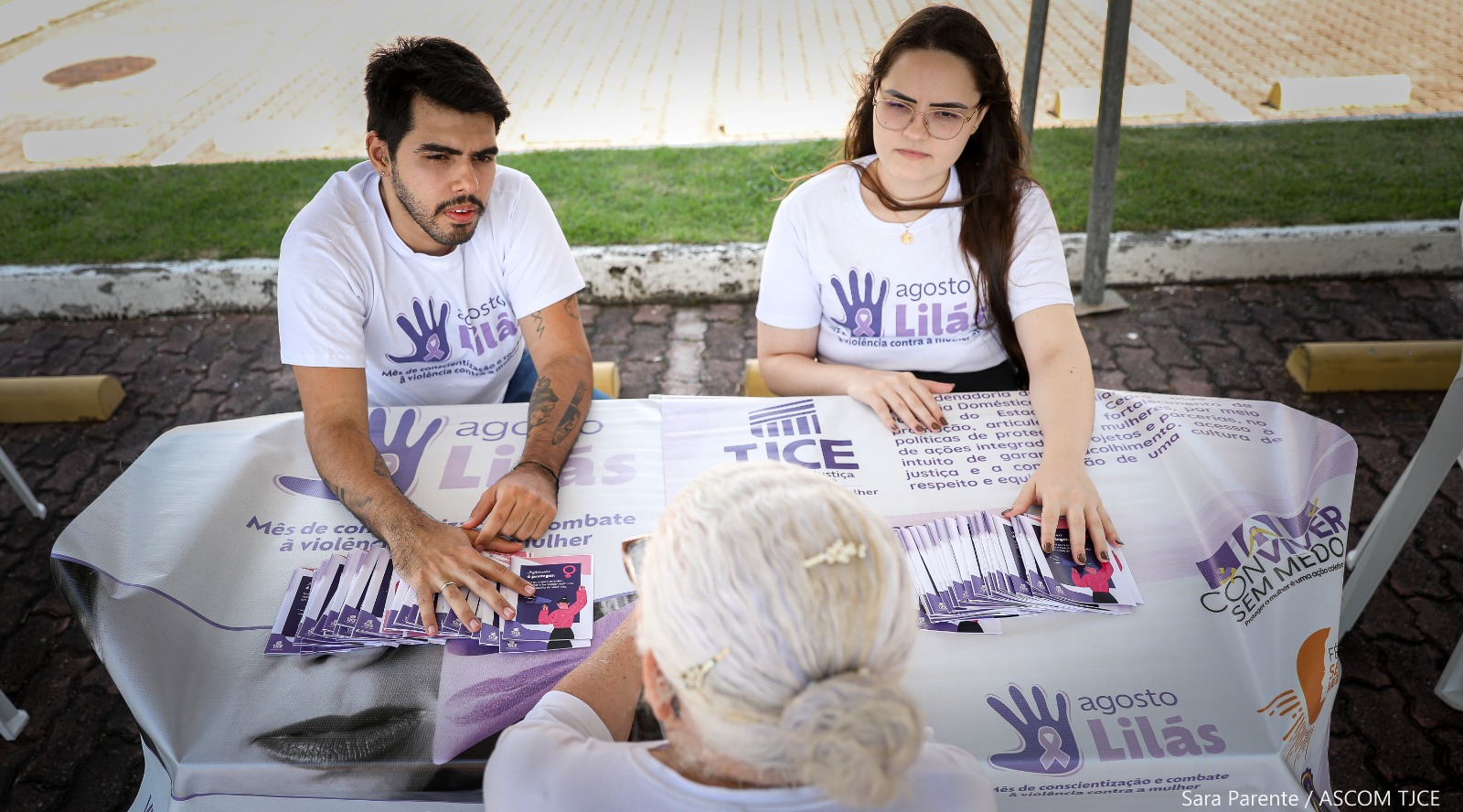 Vista superior de um jovem e uma jovem, vestindo camisetas alusivas ao Agosto Lilás, mostrando folhetos sobre violência doméstica a uma senhora.