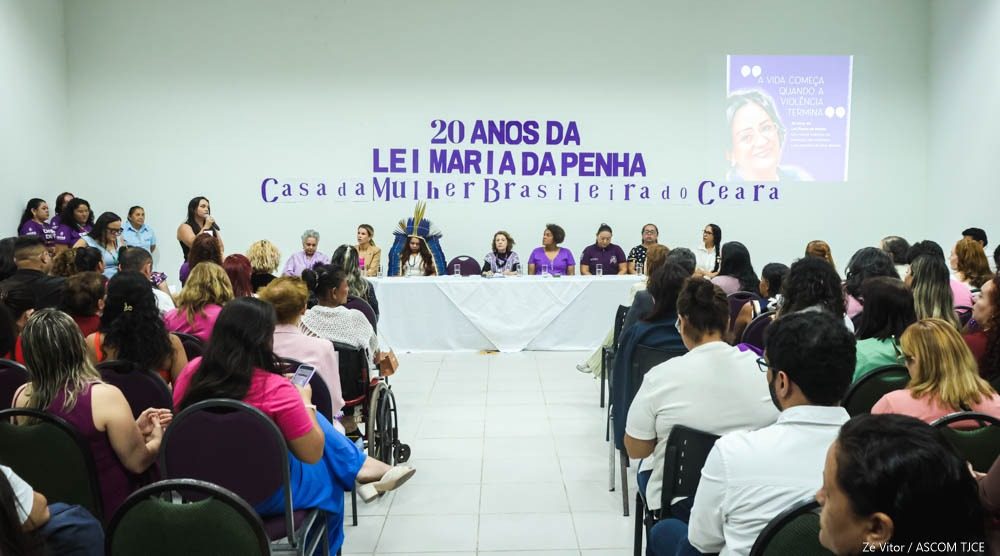 Foto geral do evento, em que se vê uma mesa com várias pessoas sentadas e, à frente delas, o púbico sentado ouvindo as palestras