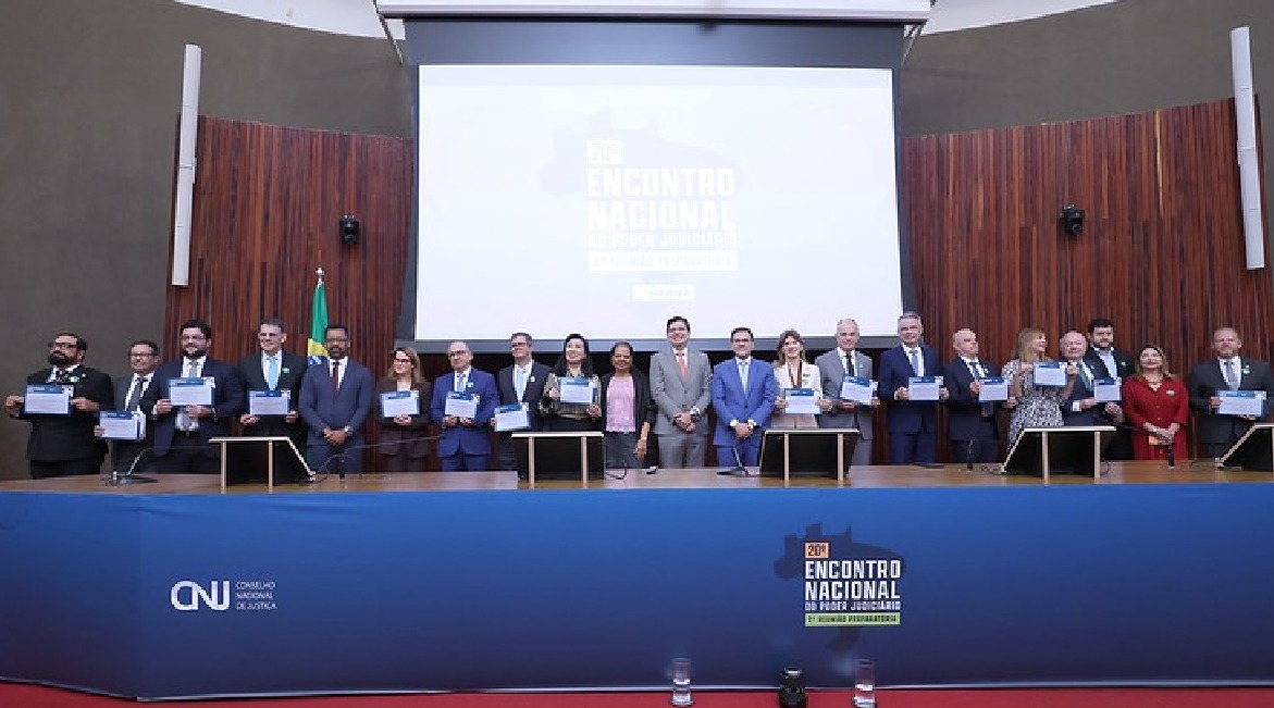 Imagem de vários homens e mulheres posando para foto, em pé, sorrindo, em um grande auditório