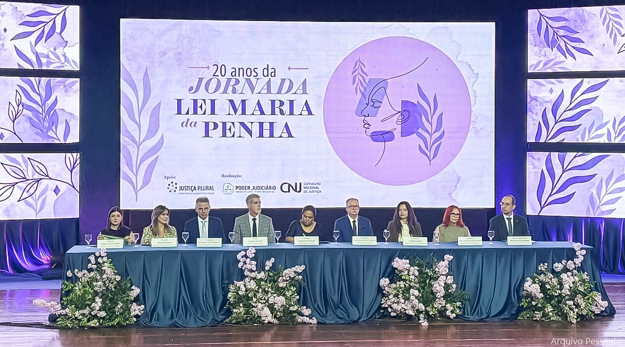 Fotografia de pessoas sentadas em uma mesa de debates com painel alusivo ao tema do evento atrás.